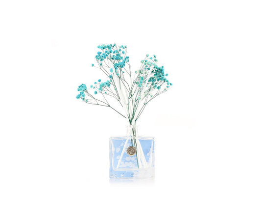 A glass bottle with a blue background and white reeds and dried blue gypsophila coming out of the top
