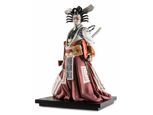A porcelain figure standing on a dark wood base. The figure is dressed in red and white Kabuki robes, with an elaborate headdress and bold make up, completed with a sword strapped to their back.