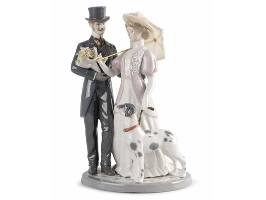 A porcelain figure of a couple in vintage dress walking their dog. The woman wears a lacy white dress and carries a parasol, while the man wears a dark grey suit and holds a bunch of flowers. It has a porcelain base.
