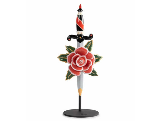 A porcelain sculpture of a dagger with an ornate handle plunging through a red rose with green leaves with a white matte blade. It has a black metal stand.