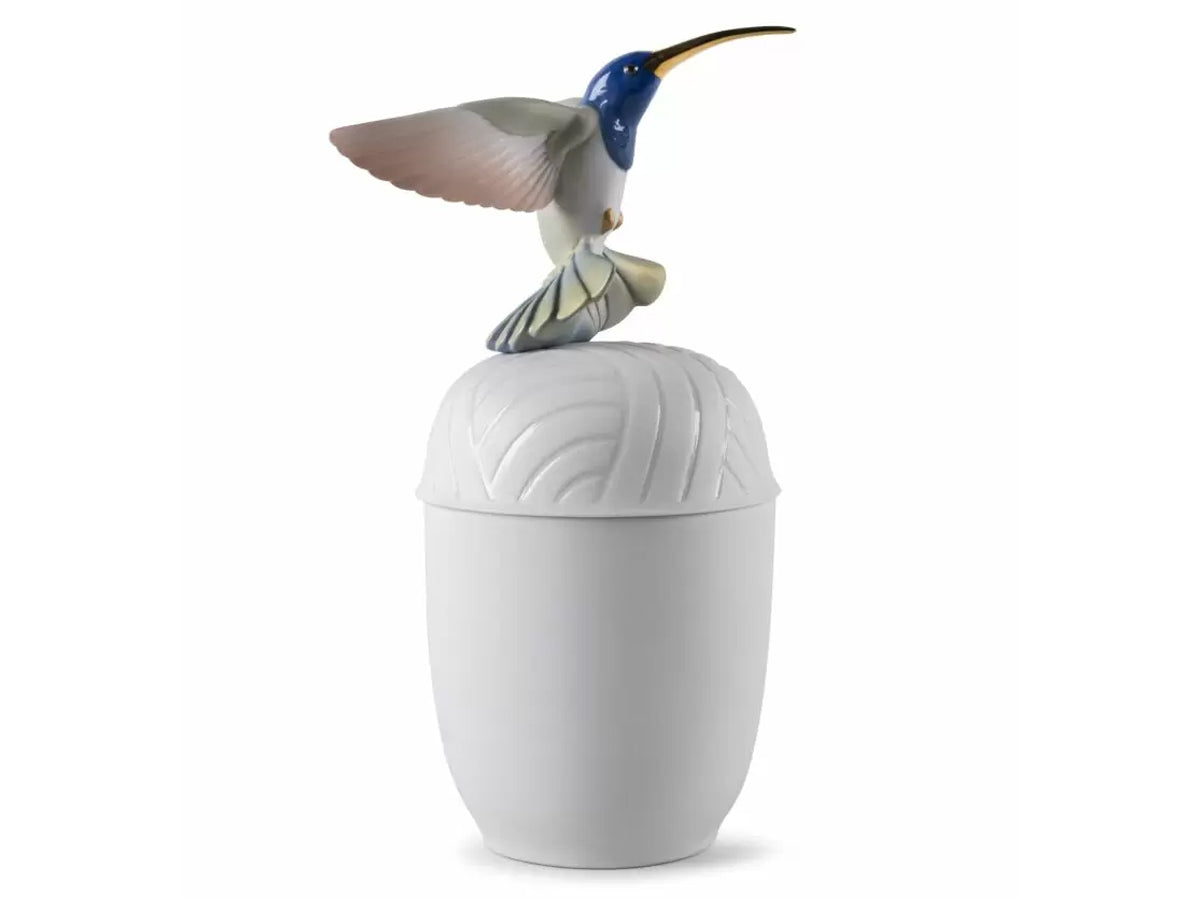 A tall matte white porcelain box with a ribbed lid, topped by a birghtly coloured hummingbird sculpture that is captured in mid-flight