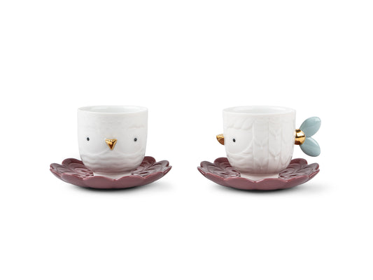 A pair of teacups and saucers. The cups are white with small bird faces and blue feathers acting as handles. The saucers are a deep reddish purple in petal patterns