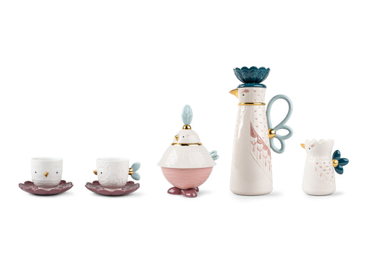 A set of two teacup and saucers, a sugar pot with a lid, a lidded coffee pot and a milk jug, all styled like quirky abstract birds in white, pink, purple and blue tones with heart details on the feathers and touches of gold luster around the handles and beaks