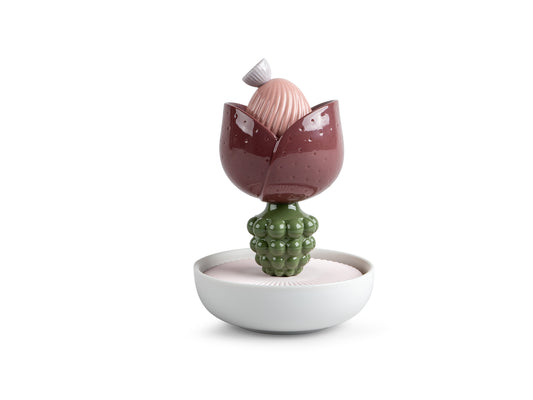 A mauve and pink abstract flower made of porcelain with a lumpy green stem sitting on a white bowl base with porous porcelain filling the bottom