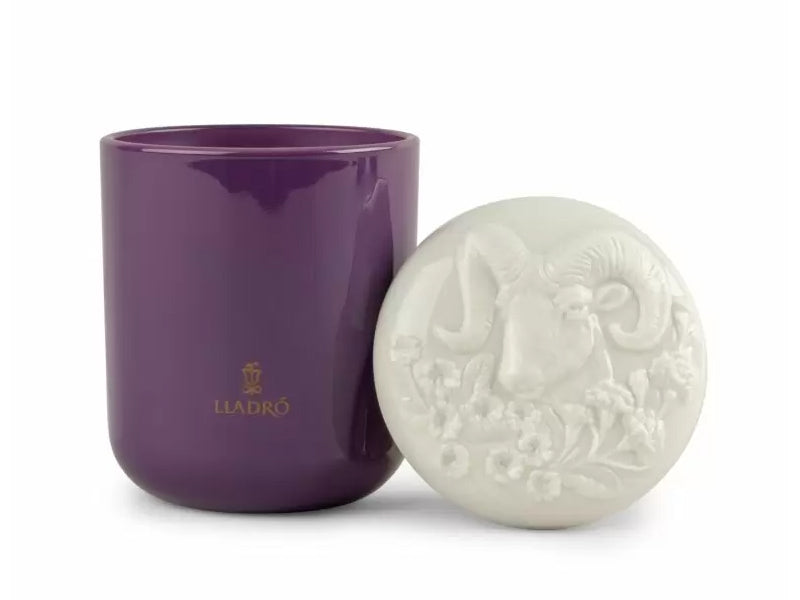 A dark purple glass candle with a white porcelain lid that is engraved with a floral pattern and a ram's head