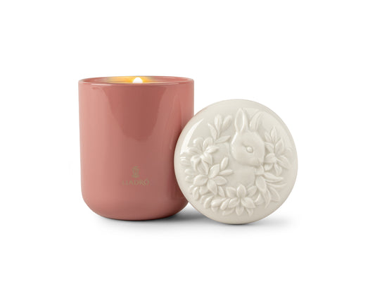 A pink glass candle with a white porcelain lid with a rabbit amongst some flowers engraved on it