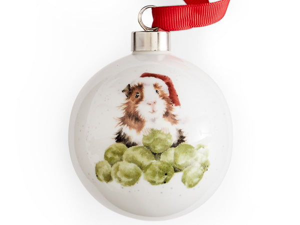 A white porcelain bauble with a silver fixture and bright red ribbon to hang it. Adorned with a watercolour illustration of a guinea pig wearing a fluffy Santa hat in the classic red and white colourway. The guinea pig is happily munching through a pile of bright green sprouts. Its tummy and mouth are a pristine white, accented by a black stripe around the belly, while its adorable face is encircled by brown fur.