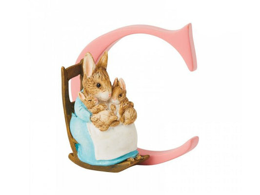 A pastel pink letter C with a statue of rabbits in a rocking chair, with a mother rabbit holding her two babies while wearing a blue dress