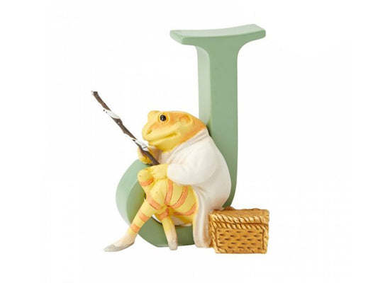 A sage green capital J with a yellow and orange striped frog sitting in the curved bottom, carrying a fishing rod and sitting next to a picnic basket.