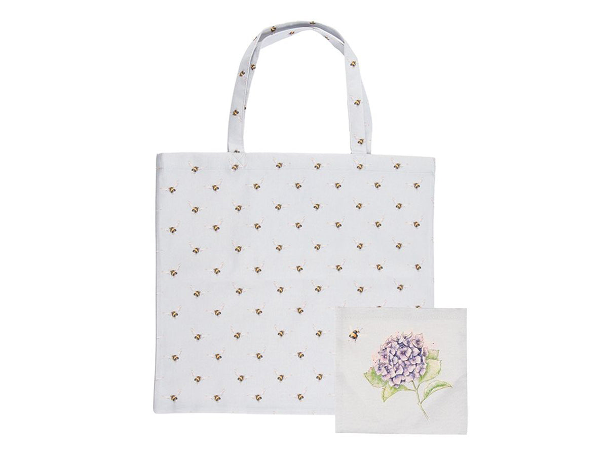 A foldable, light blue, cotton tote bag with bees on it, and a purple hydrangea flower on the pocket inside.