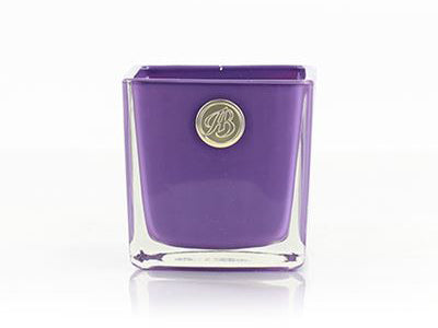A purple soy wax candle in a square glass votive with a gold logo on one side.