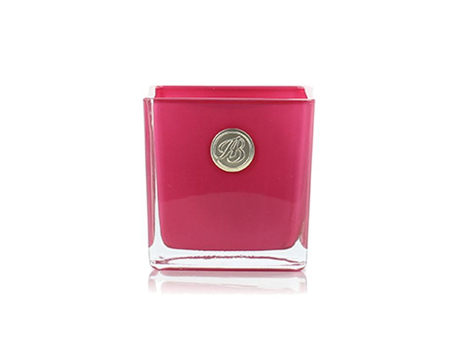 A pink candle in a square glass votive with a gold logo on the front