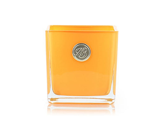 An square orange glass votive with a candle inside, complete with a round gold logo on the front