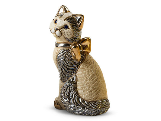 A porcelain figurine of a cat sitting on its hide legs, the fur is cream with grey accents, finished in a pearlescent enamel. The cat has a golden bow around its neck.