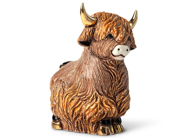The image shows a porcelain Highland cow figurine set against a plain light background. The cow is seated upright with its legs tucked beneath its body. It features a textured, flowing coat rendered in warm copper and bronze tones with subtle golden highlights that accentuate the sculpted fur details.

The cow’s curved horns are painted in a glossy gold finish, contrasting with its soft cream-colored muzzle. Its expressive dark eyes peek out from beneath a long fringe of sculpted hair that drapes over its f