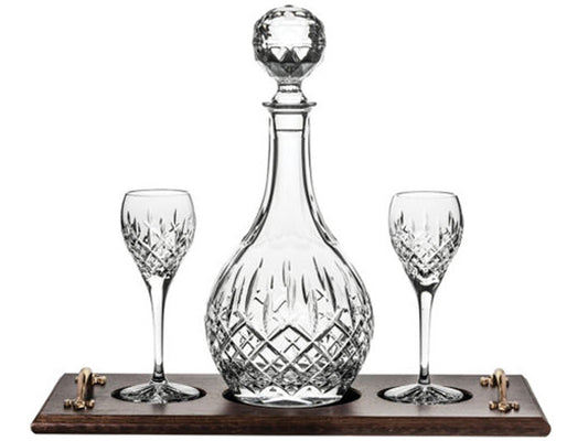 A crystal port decanter and matching stemmed glasses with a cut pattern on the outside, sitting on an oak tray