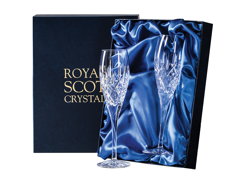 A pair of cut crystal champagne flutes with slender stems, featuring a bed of diamonds at the base of the flute and single dashes pointing up towards the smooth rim, stored in a navy blue silk-lined presentation box