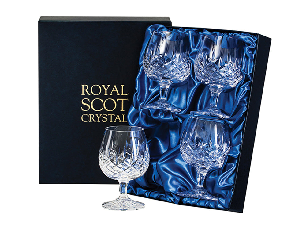 A boxed set of four crystal brandy glasses with rounded bowls and short stems, envrgaved with a bed of diamonds at the base of the bowl and single dashes reaching up towards the smooth rim of each. The box is silk-lined and navy blue, embossed with Royal Scot's branding in gold lettering on the lid