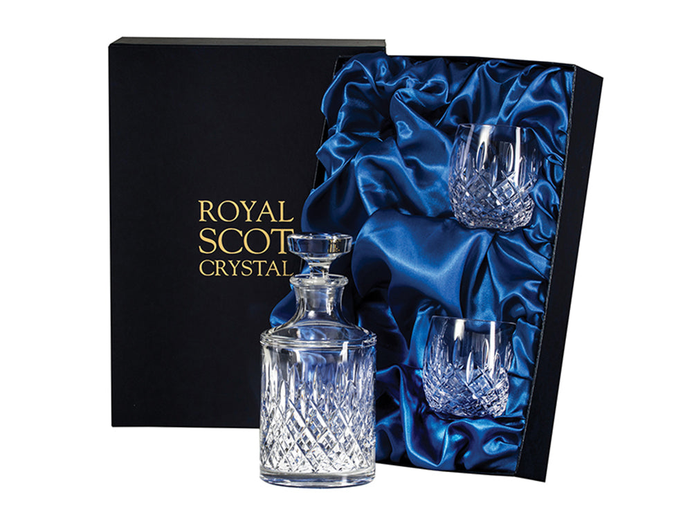 A set of two barrel-shaped tumblers and a round spirit decanter with a flat stopper in a navy-blue silk-lined presentation box with gold branding on the lid. The pieces are engraved with a bed of diamonds and vertical flicks