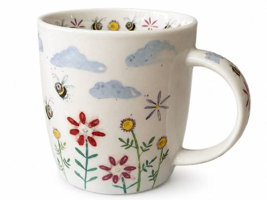 A white ceramic mug decorated with a whimsical pattern of blue clouds, yellow and red flowers, green stems, and bumblebees, shown against a plain white background.
