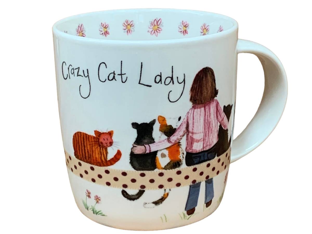 A white ceramic mug featuring a whimsical illustration of a woman from behind petting several cats, with the text 'Crazy Cat Lady' written above. The mug is decorated with pink flowers around the rim and a polka-dotted band near the bottom.