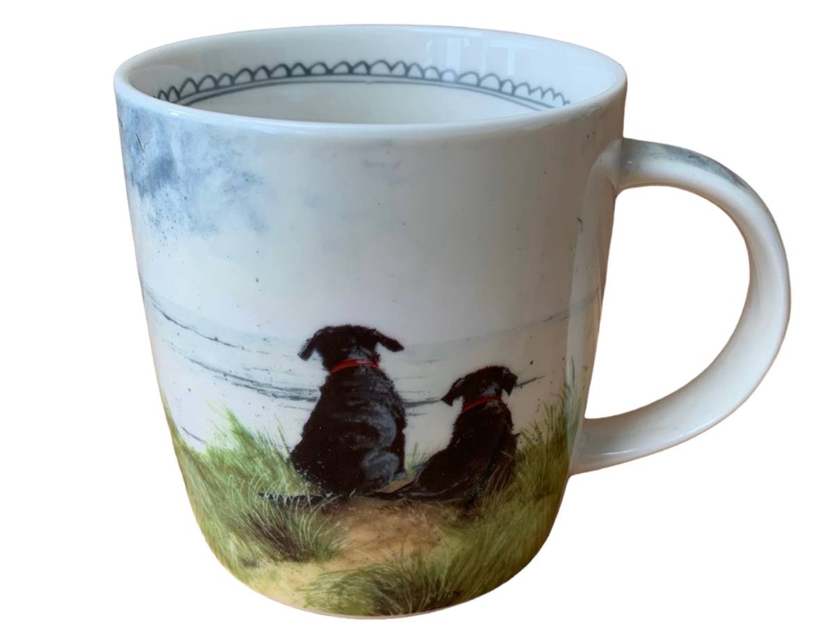 A white ceramic mug featuring a hand-painted illustration of two black dogs sitting in grassy dunes by the sea, with a blue patterned rim.