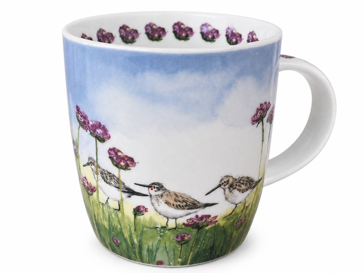 A white ceramic mug decorated with a watercolor-style illustration of three sandpipers standing in a grassy field with purple flowers under a blue sky.
