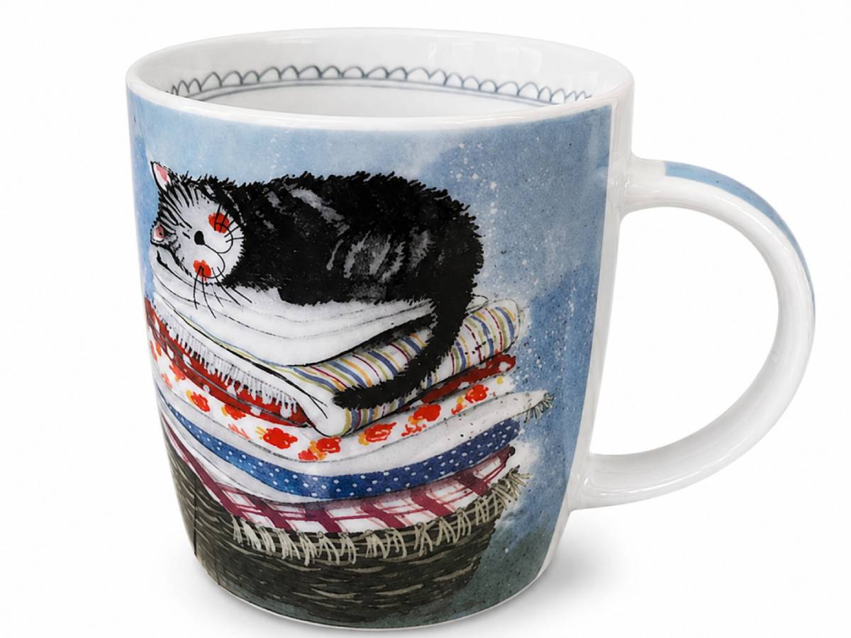 A white ceramic mug featuring a whimsical illustration of a black and white cat sleeping on top of a stack of patterned quilts against a light blue background.