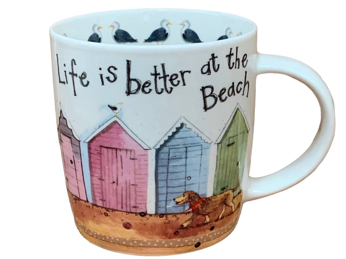 A white ceramic mug featuring a beach-themed illustration with the text 'Life is better at the Beach'. The design includes colorful beach huts in pink, blue, and green, a brown dog, and seagulls along the inner rim, set against a plain white background.