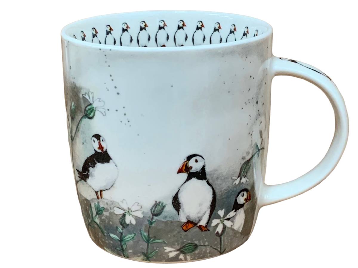 A white ceramic mug featuring a printed illustration of puffins and white flowers, with a row of puffin figures around the inner rim, shown against a plain white background.