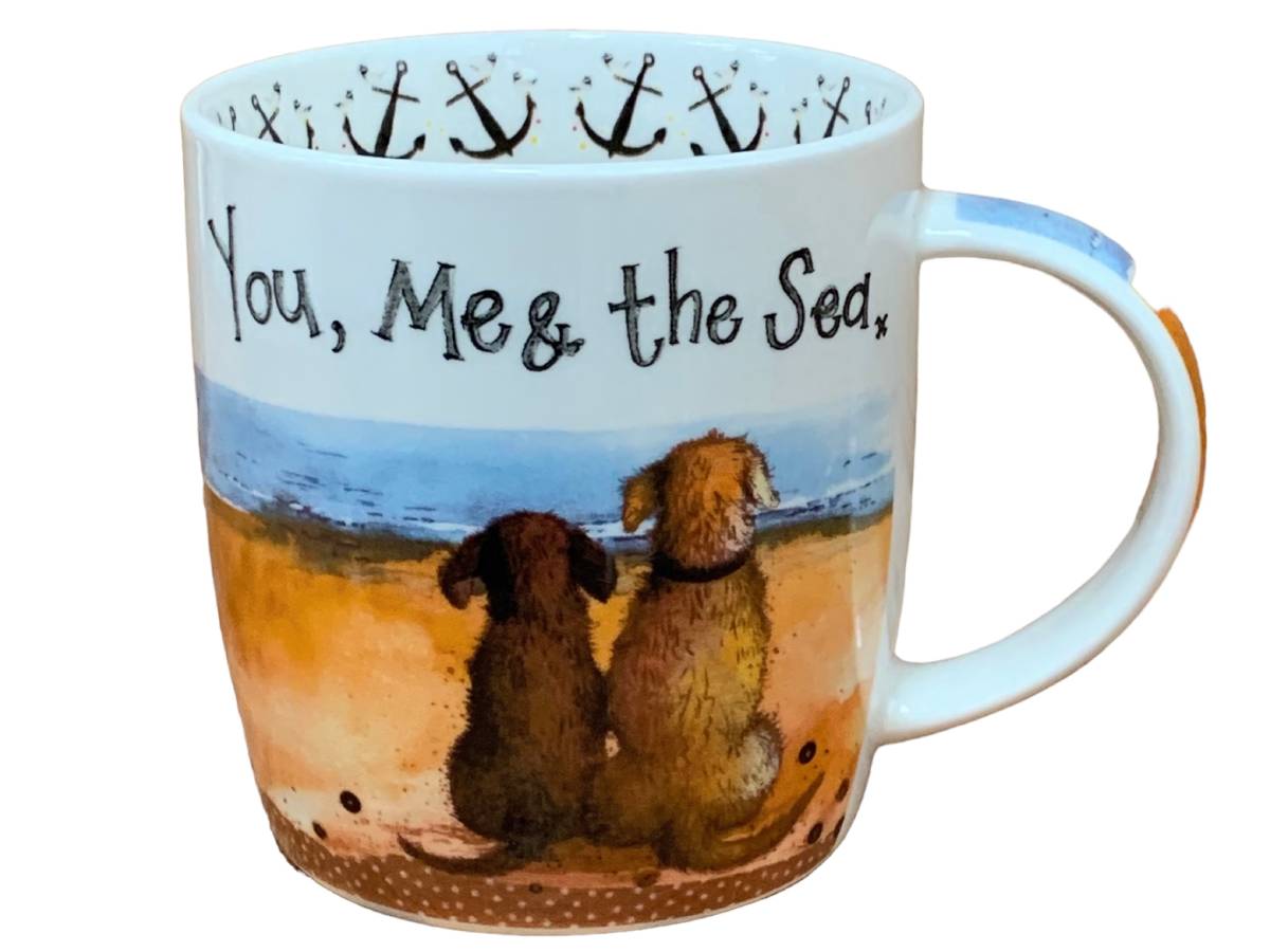 A white ceramic mug featuring a printed illustration of two dogs sitting on a beach facing the sea, with the text 'You, Me & the Sea' on the front. The inside rim is decorated with black anchors, and the handle has a light blue accent.