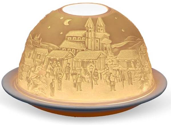 A dome-shaped decorative lamp or light fixture with an embossed winter village scene on the inside, glowing warm yellow light. The design features a church, houses, trees, and small figures of people under a crescent moon and stars.