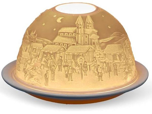 A dome-shaped decorative lamp or light fixture with an embossed winter village scene on the inside, glowing warm yellow light. The design features a church, houses, trees, and small figures of people under a crescent moon and stars.