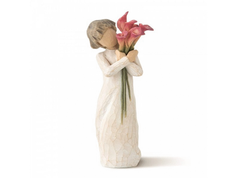 A faceless cast stone figurine: feminine figure painted in soft earthy tones. She wears a creamy white crackled dress and sports soft brown hair, cut into a bob. She is embracing a bouquet of pink calla with contrasting greens stems. The face is featureless for personal interpretation.