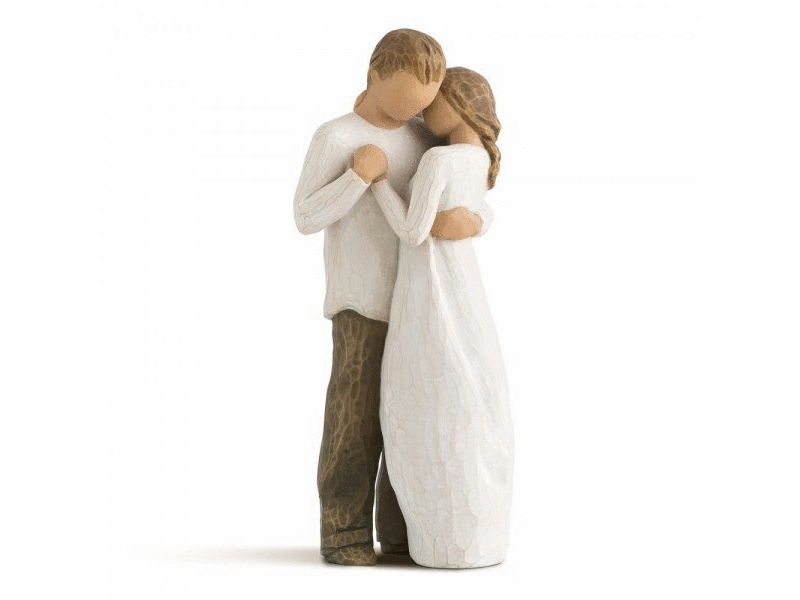 A cast stone figurine by Willow Tree, depicting a faceless couple holding each other close. The masculine figurine holds the hand of the feminine figurine close to his chest, whilst they both wrap their remaining hand behind one another's back. Painted in earthy, soft tones of cream, grey, beige and brown.