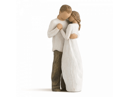 A cast stone figurine by Willow Tree, depicting a faceless couple holding each other close. The masculine figurine holds the hand of the feminine figurine close to his chest, whilst they both wrap their remaining hand behind one another's back. Painted in earthy, soft tones of cream, grey, beige and brown.