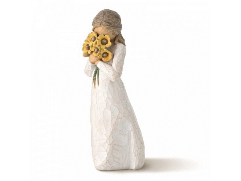 This Willow Tree figurine is made of cast stone and features a faceless girl holding a bouquet of yellow flowers close to her chest and face. Painted in earthy tones with a white dress, brown hair, and beige skin.