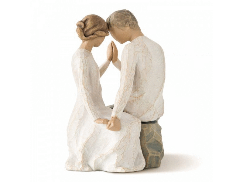 A cast stone sculpture shows a couple, a male and a female, seated on a stone, gently touching foreheads and intertwining their hands, which are raised and touching palms. The figurine is painted in natural, earthy hues, featuring white and grey attire. Notably, their facial features are intentionally absent, to allow for personal interpretation.