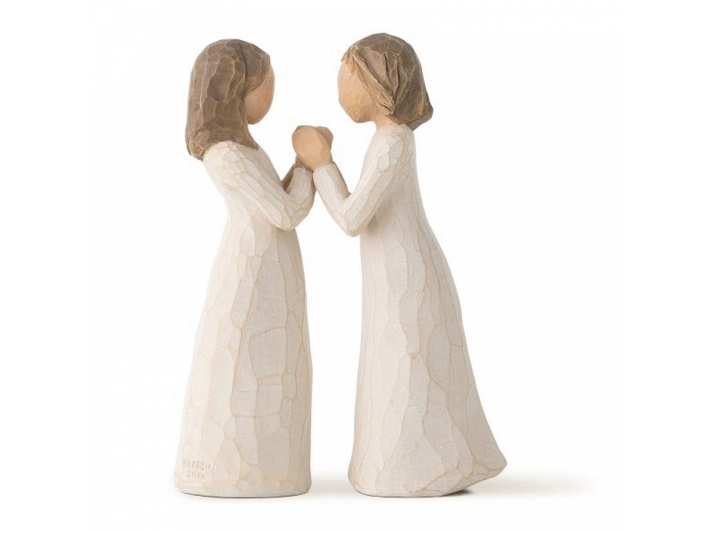 A duo of faceless feminine figurines with white gowns stand across from one another, clasping each others hands and slightly leaning towards one another. They are painted in a muted colour palette of soft, earthy tones.