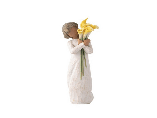 A cast stone figurine of a featureless feminine figure with light terracotta skin. She wears a white dress with a crackle texture, exposing the natural brown hues of the stone underneath. Clasped in her hands is a vibrant bundle of yellow calla lilies, beautifully contrasting with the soft earthy tones of the figurine's paint palette.