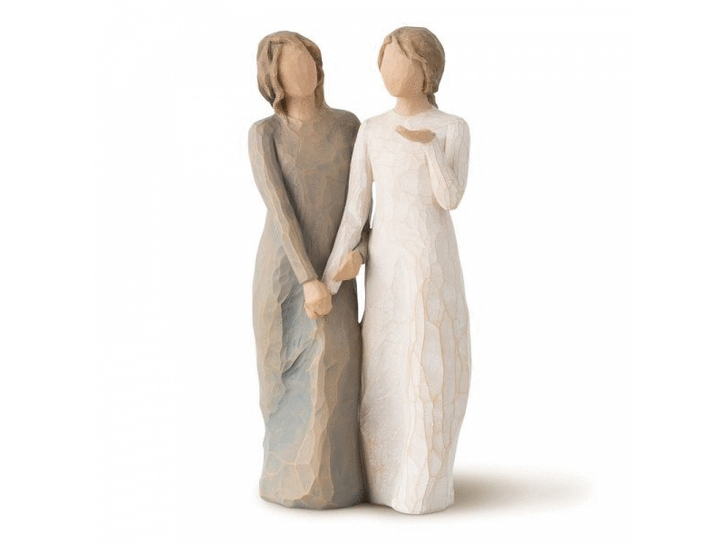 A cast stone figurine of two older sisters with featureless faces. One sister wears a cool toned brown dress with light brown hair and the other sister wears a white dress with light brown hair. The two sisters are holding hands, the sister in the dark dress holds both the hand and arm of the white dress sister. The sister in the white dress has her free hand held up with palm facing out.