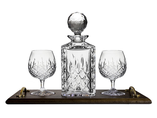 A crystal decanter and two matching brandy glasses with a cut pattern on the outside, sitting on a handled oak tray