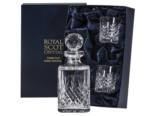 A crystal whiskey set including a square spirit decanter and two whiskey tumblers, all featuring a triple-flick pattern that's cut into the lower portions of each piece. It comes in a navy blue silk-lined presentation box with the brand's name and information embossed in gold on the front of the lid.