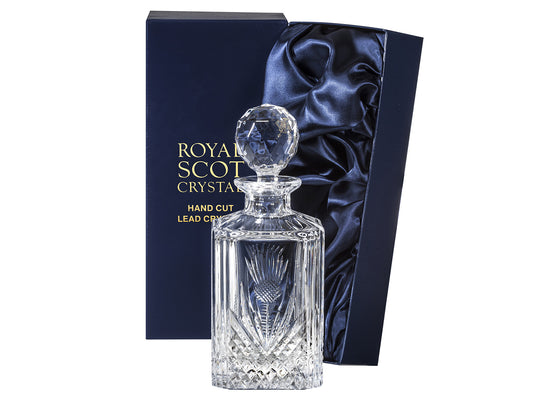 A square crystal decanter that is cut with a bed of diamonds around the base with a Scottish Thistle on each side, with a smooth top and a golf-ball style topper. It comes in a navy-blue silk-lined presentation box with gold branding on the lid.