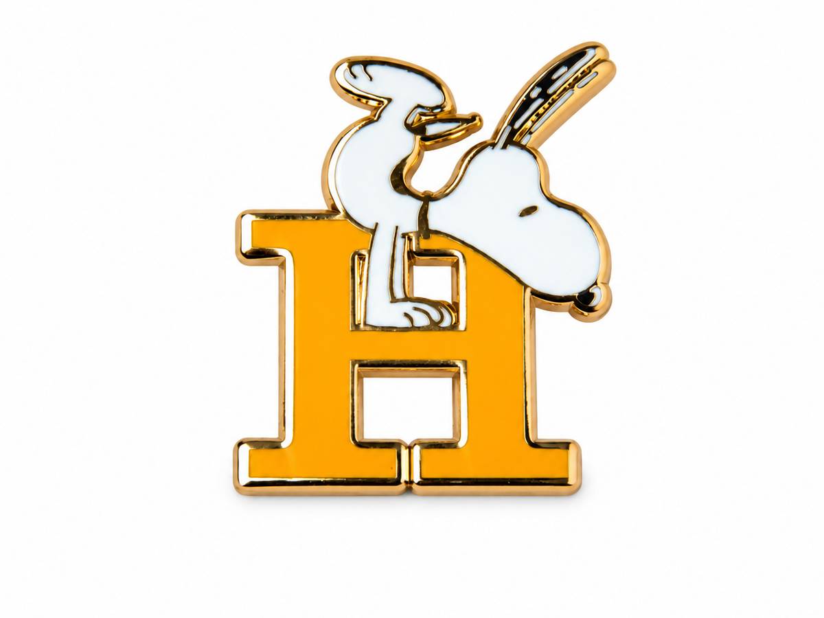 A gold and white enamel pin featuring the character Snoopy sitting on a large letter H.