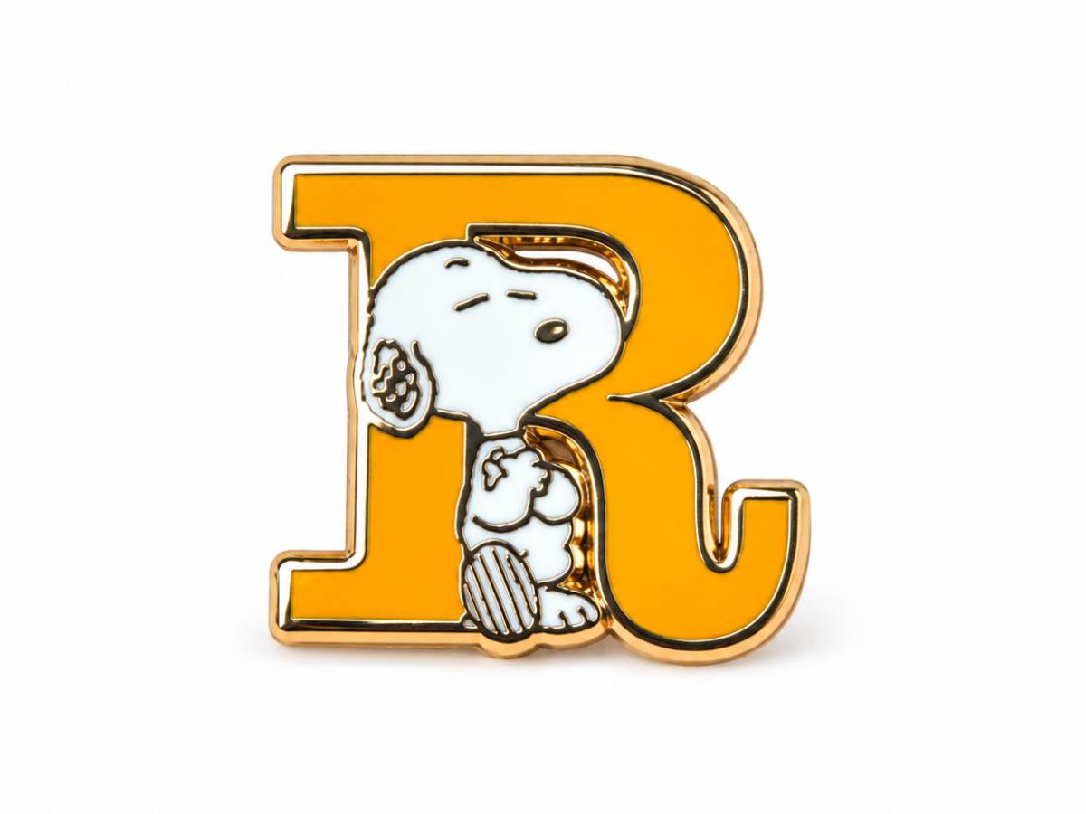 An enamel pin featuring the letter 'R' in bright yellow with a gold-tone outline, and Snoopy sitting inside it in white with black details.
