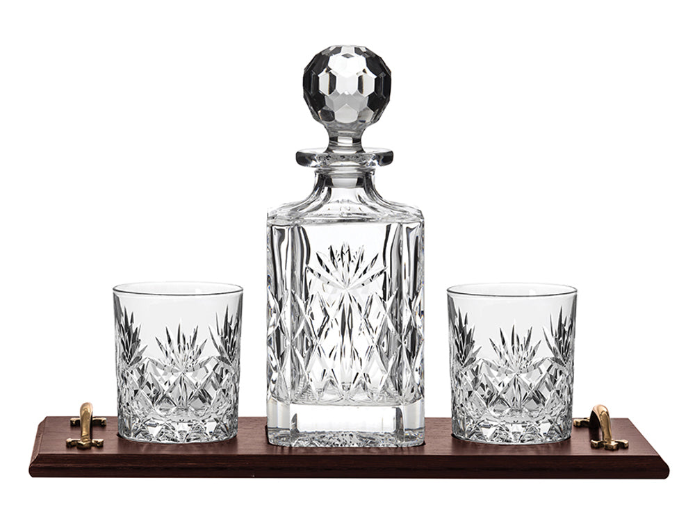 A crystal decanter and two glasses with a cut design on the outside, on an oak tray with gold handles